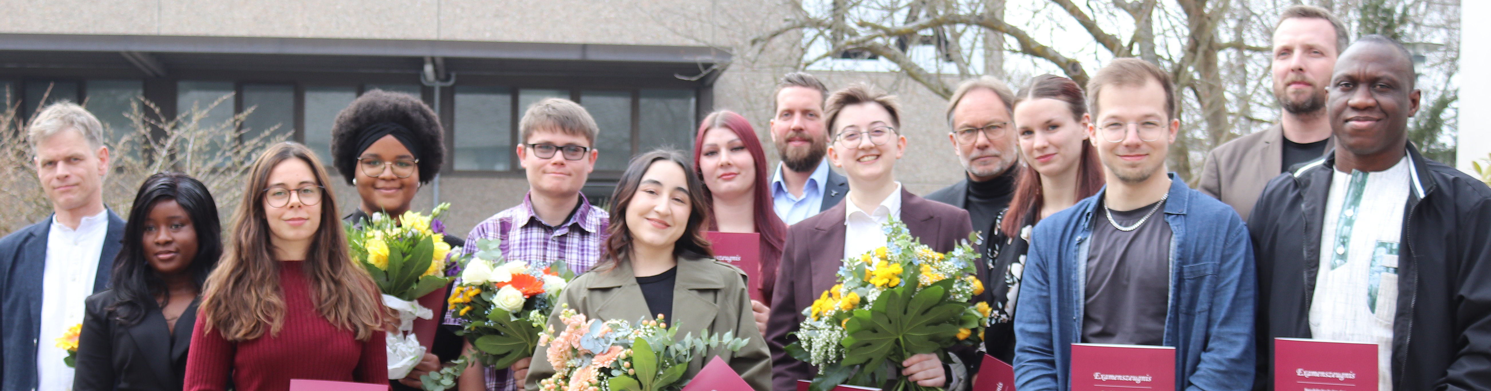 Auf dem Foto stehen junge Männer und Frauen, die ihre Examensurkunde bekommen haben. Die besten Absolventen halten Blumen in den Händen