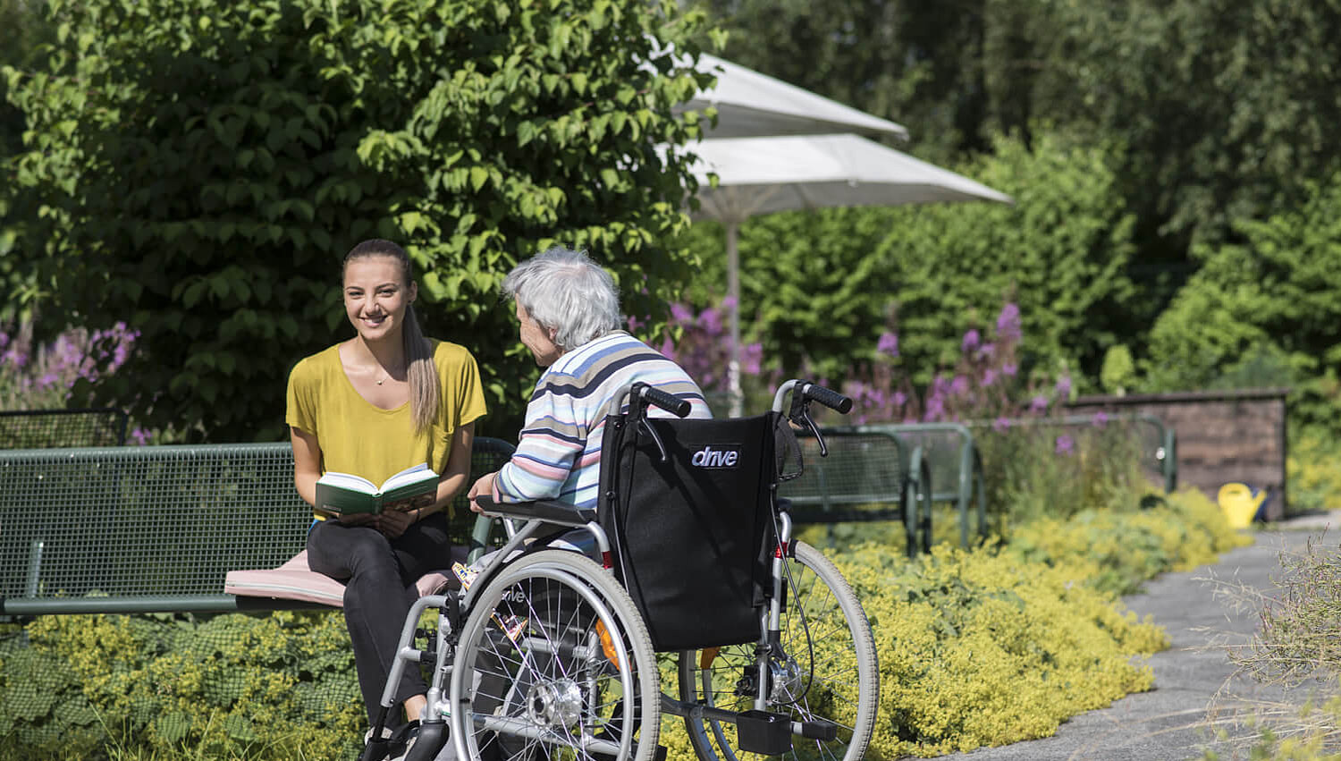 Junge Frau liest älterer Dame im Rollstuhl im Garten der Bezirksklinik Rehau ein Buch vor.